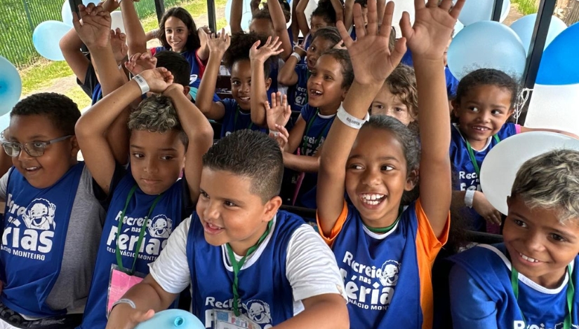 Grupo de crianças com coletes azuis sorriem com mãos levantadas