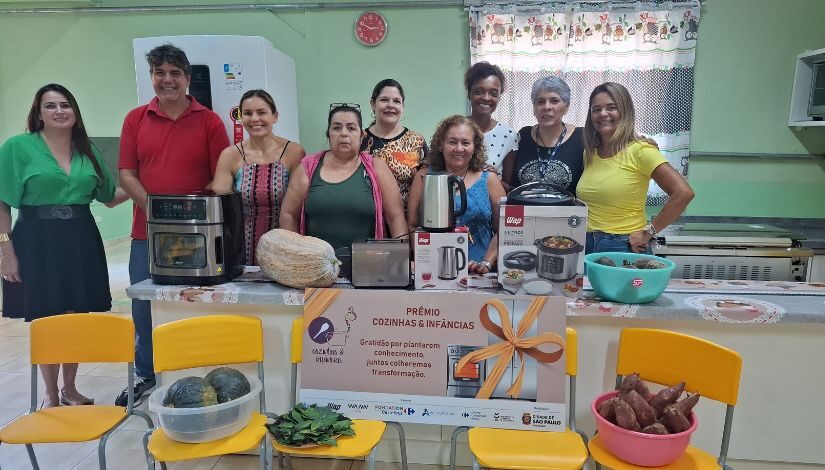 Fotografia mostra grupo de nove pessoas, sendo 8 mulheres e 1 homem, posando para foto. Na frente deles, há uma bancada com eletrodomésticos.