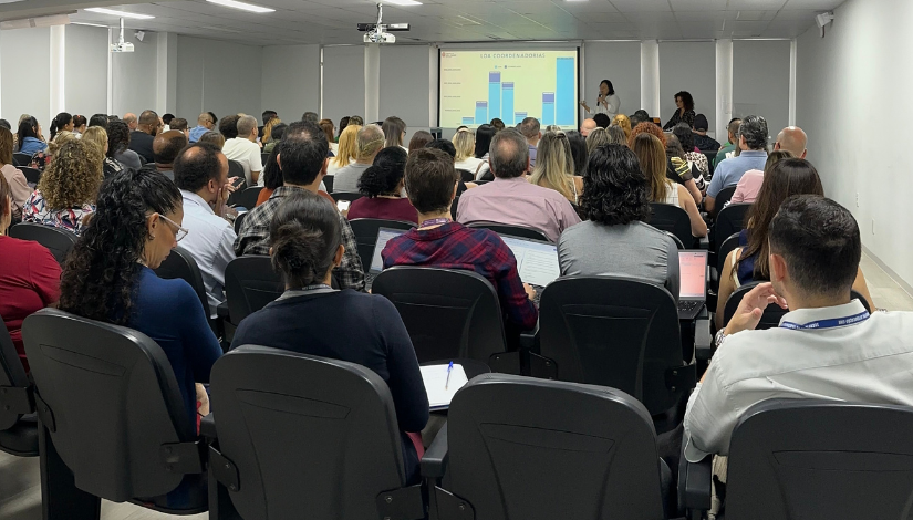 Pessoas sentadas de costas para a foto enquanto prestam atenção no gráfico que está na tela.