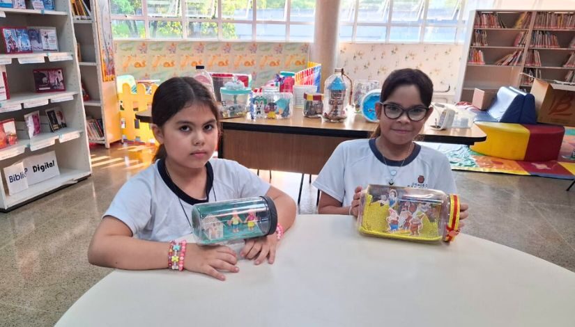 Duas meninas estão sentadas, cada uma segura um pote que está na mesa à frente delas.