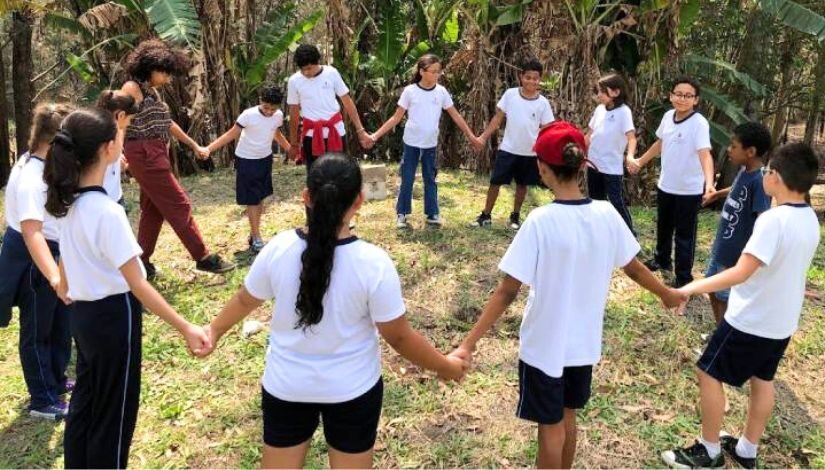 Fotografia mostra estudantes em uma área verde, eles estão de mãos dadas em círculo. Os alunos são da EMEF Philó Gonçalves dos Santos e fazem parte do projeto 