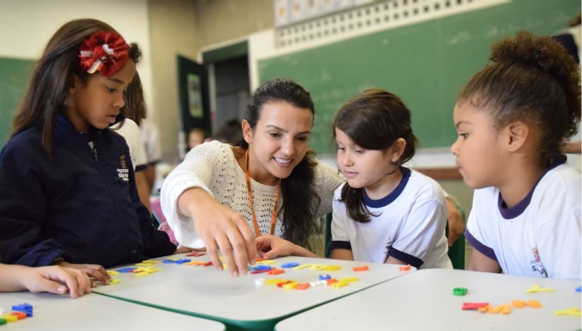 Fotografia mostra professora sentada com três estudantes ao seu lado. Na mesa há frente delas há peças de montar.