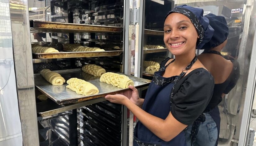 Imagem com uma mulher com avental azul sorrindo para a câmera enquanto segura uma forma de pão