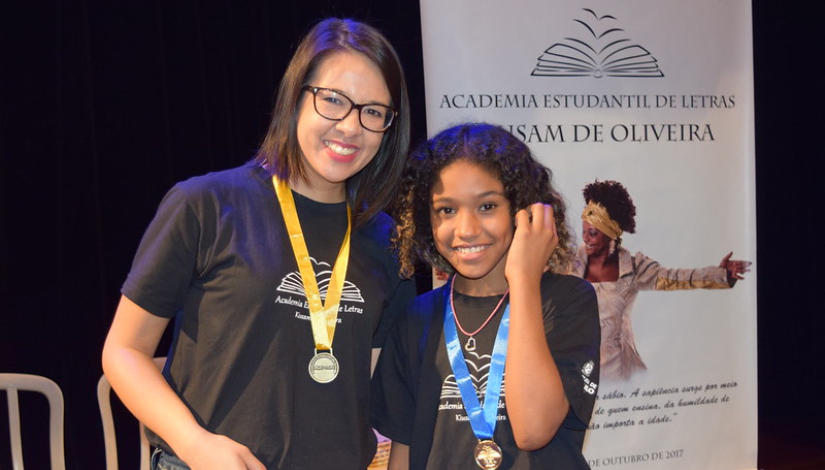 Uma mulher e uma adolescente sorriem para a câmera. Ambas vestem camisetas pretas com o logotipo da AEL e utilizam medalhas no pescoço — uma com fita dourada e a outra com fita azul. Ao fundo, há um banner da AEL com o nome