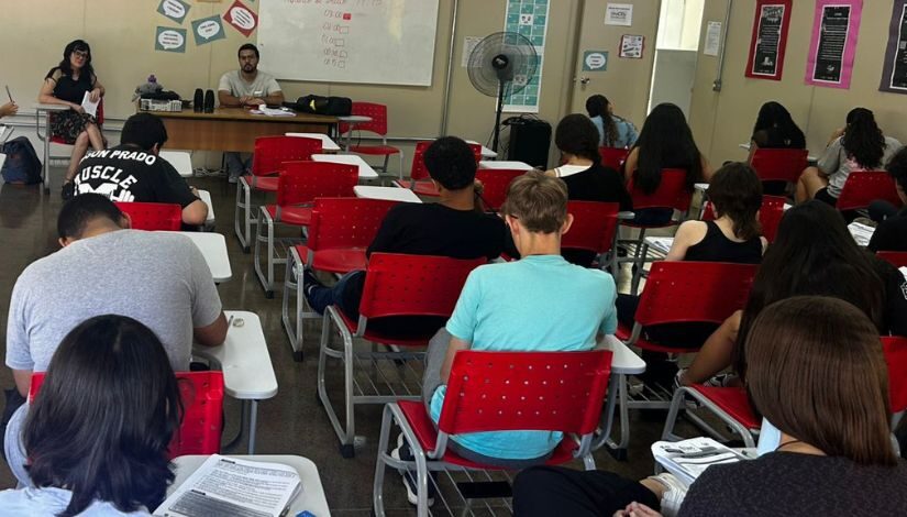 Fotografia de estudantes em sala de aula. Ao fundo dois professores.