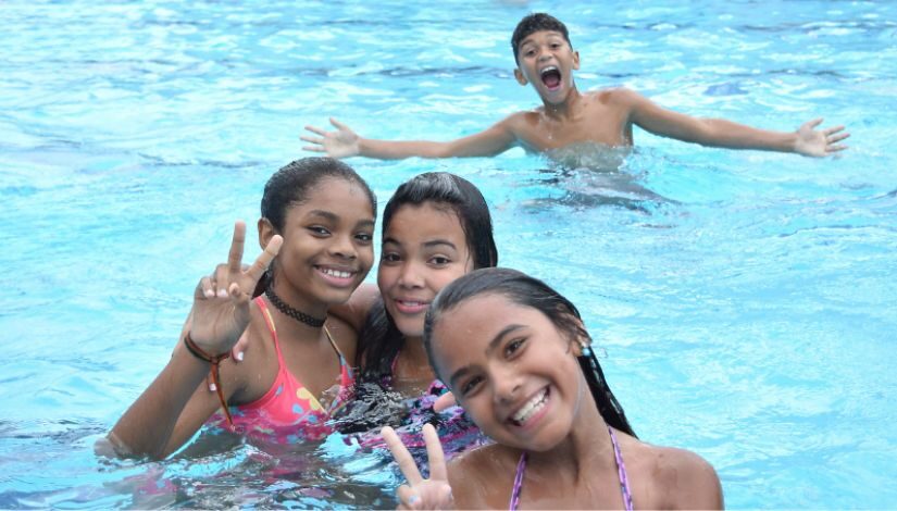 Na imagem, quatro crianças se divertem em uma piscina. Três meninas estão mais próximas da câmera, sorrindo. Todas estão com roupas de banho coloridas e molhadas. Ao fundo, um menino também está na água, com os braços abertos e expressão animada.
