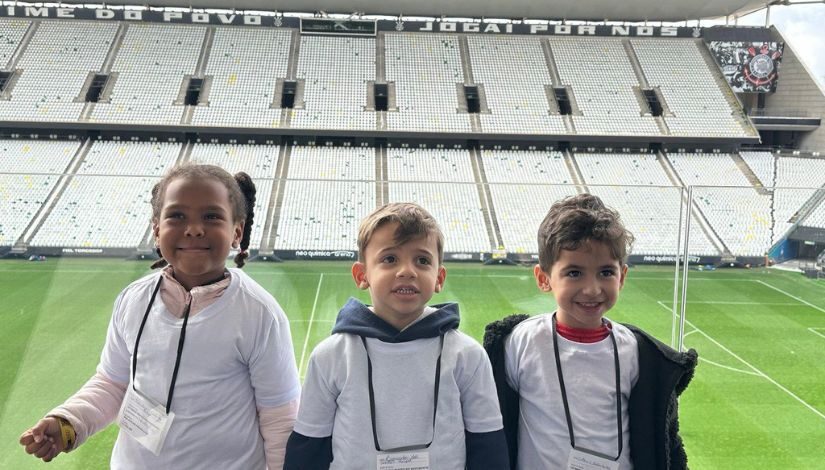 Na imagem, vemos três crianças pequenas, do CEI Maria Beatriz Nascimento, sorrindo e posando para a foto dentro da Arena Corinthians, um famoso estádio de futebol em São Paulo. Elas estão usando crachás e camisetas brancas. Ao fundo, aparecem as arquibancadas vazias com cadeiras brancas.