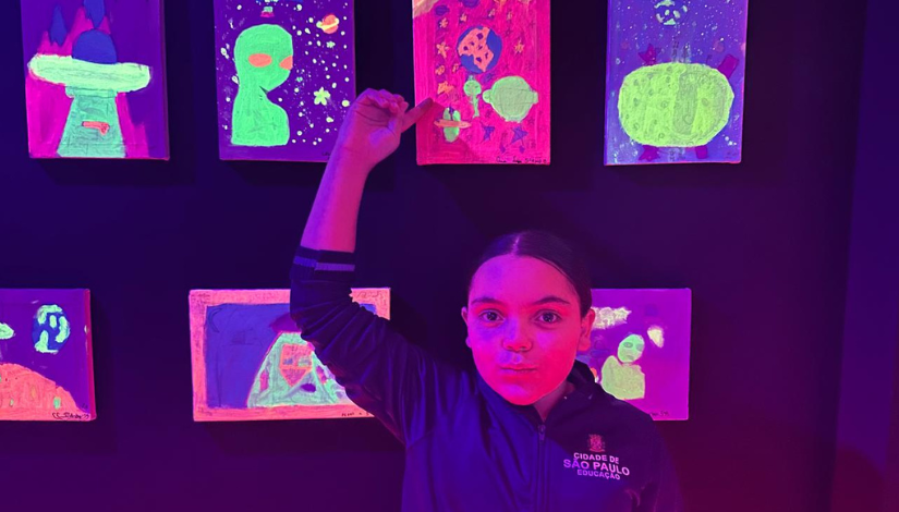 Imagem com uma estudante com uniforme da Prefeitura de São Paulo apontando para um quadro fluorescente