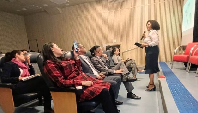 Fotografia mostra pessoas em um auditório durante o 1º Fórum sobre Racismo da DRE Butantã. A frente uma mulher negra segura o microfone e conversa com a platéria.