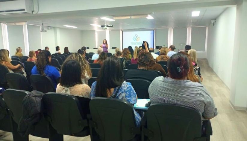 Diversas pessoas sentadas, voltadas para frente, assistindo a uma apresentação. Uma mulher está em pé, ao centro, falando diante de uma tela de projeção onde aparece o logotipo do Gabinete Integrado de Proteção Escolar.