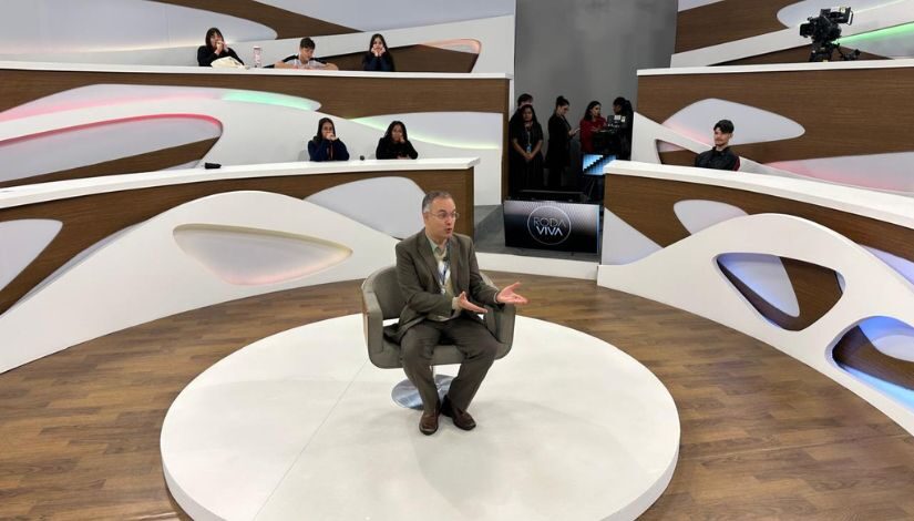 Imagem com um homem de terno em uma cadeira de um estúdio de televisão