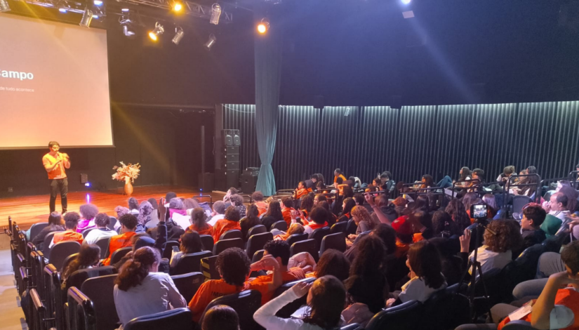 Auditório com crianças sentadas. No palco do auditório, Estevan está em pé, com camisa laranja e calça petra, segurando o microfone, ministrando o workshop
