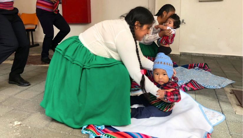 Fotografia de famílias migrantes do CEU Nova Luz onde terá oferta gratuita de serviços essenciais. No centro uma mãe boliviana com trajes típicos , saia verde e blusa branca. Ela agacha para pegar seu bebê que está sentado sobre uma manta.