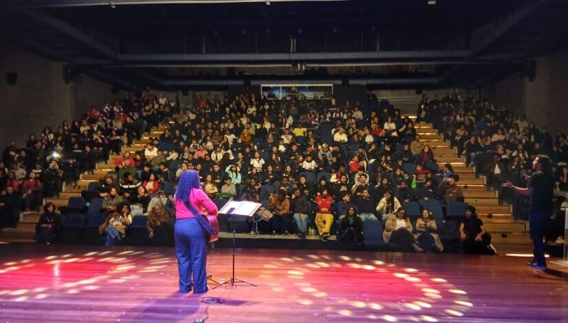 Imagem de um teatro com uma pessoa no palco tocando violão