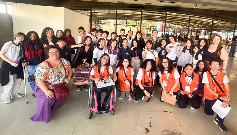 Educomunicação Inclusiva. Fotografia de uma turma de estudantes e sua professora posando para a foto. na frente um grupo de estudantes de colete laranja do Imprensa Jovem.