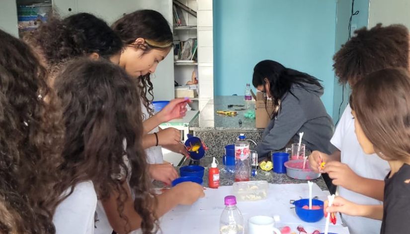 Estudantes em momento de investigação e descobertas na aula de Ciências. Várias meninas estão ao redor da mesa com potes azuis, tintas coloridas, garrafa plática e detergente.