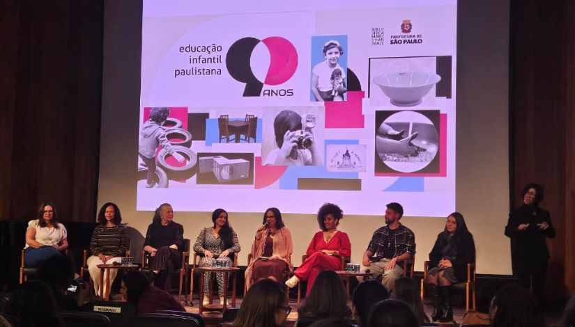 Fotografia mostra oito pessoas sentadas em cadeiras lado a lado no palco celebrando os 90 Anos Educação Infantil da Rede Municipal, na Biblioteca Mário de Andrade.