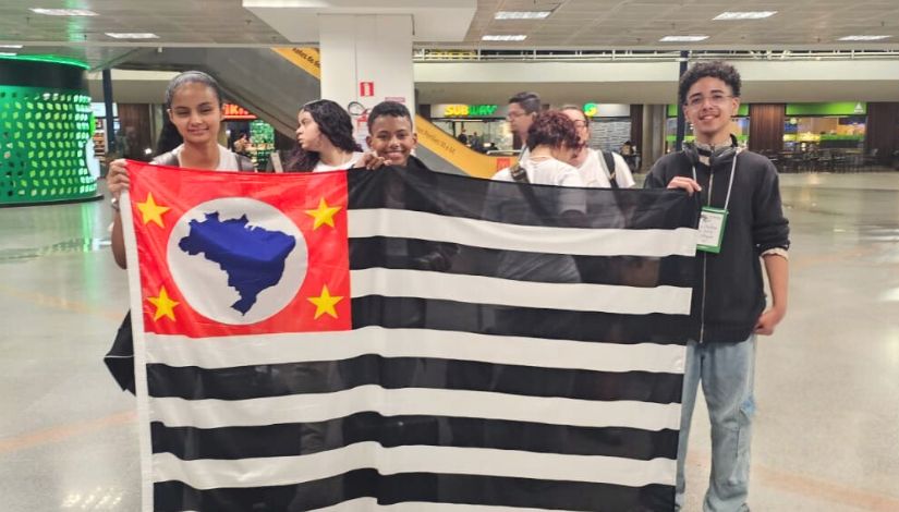 Fotografia dos estudantes Joanna Santos da Silva e Yuri Monteiro da Silva no aeroporto segurando a bandeira do Estado de São Paulo.