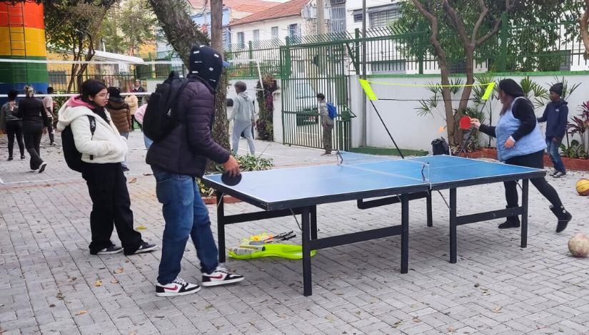 Fotografia de estudantes em área externa de escola jogando volei ao fundo e pingue-pongue em primeiro plano no