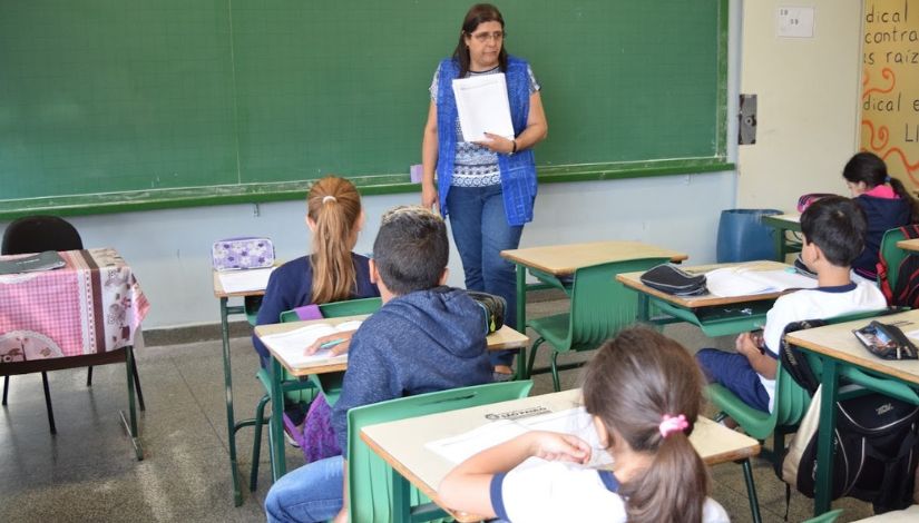 Fotografia de uma professora em pé na sala de aula em freten aos alunos que estão sentados e olhando para ela.