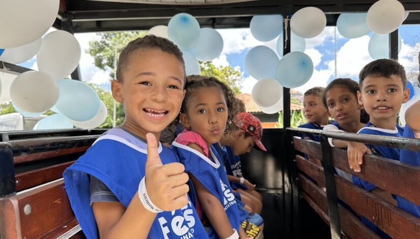 Imagem de duas crianças olhando para foto - uma está fazendo sinal de positivo e a outra está mandando um beijo - elas usam colete azul do Recreio nas Férias.