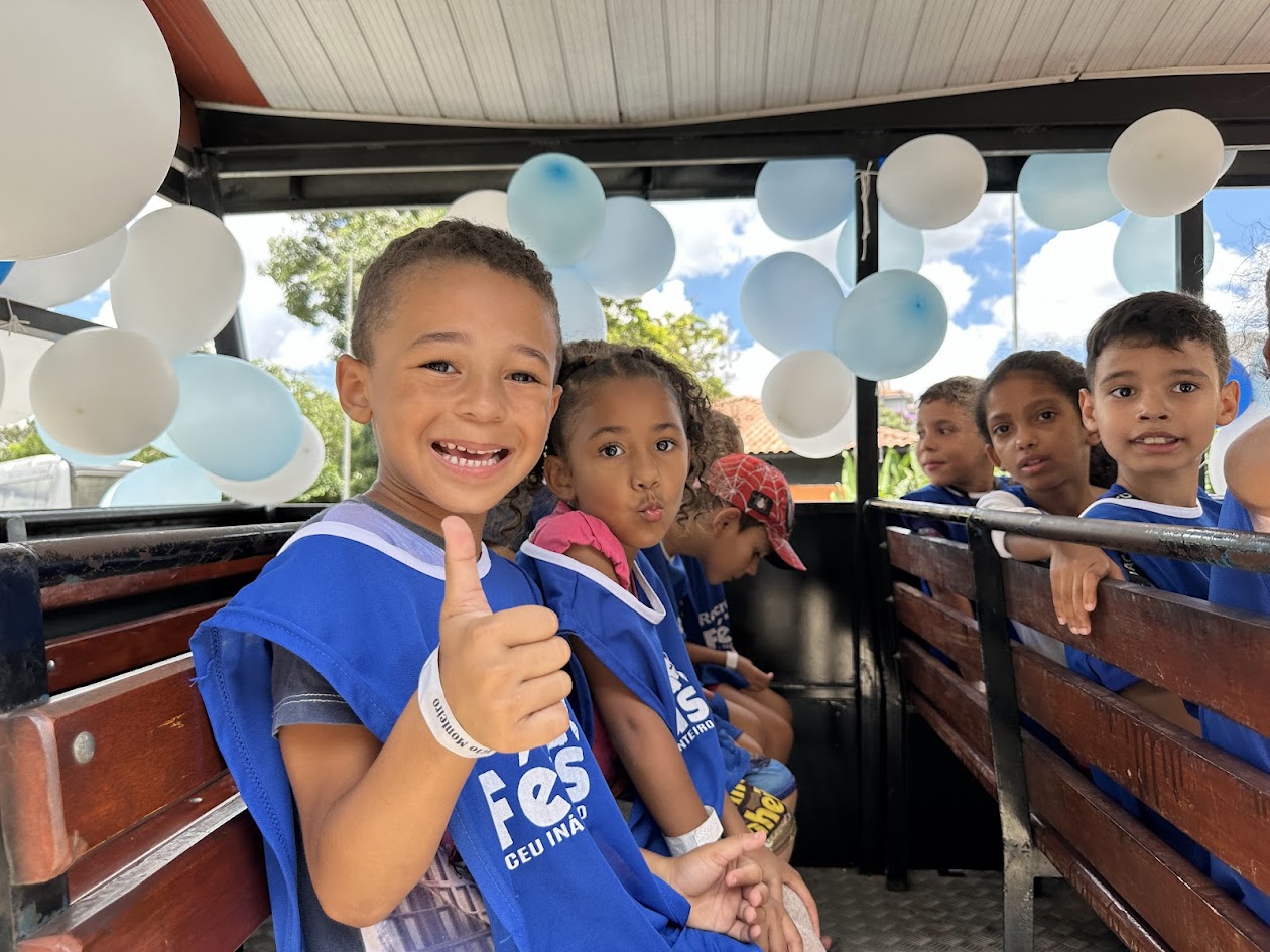 Imagem de duas crianças olhando para foto - uma está fazendo sinal de positivo e a outra está mandando um beijo - elas usam colete azul do Recreio nas Férias.