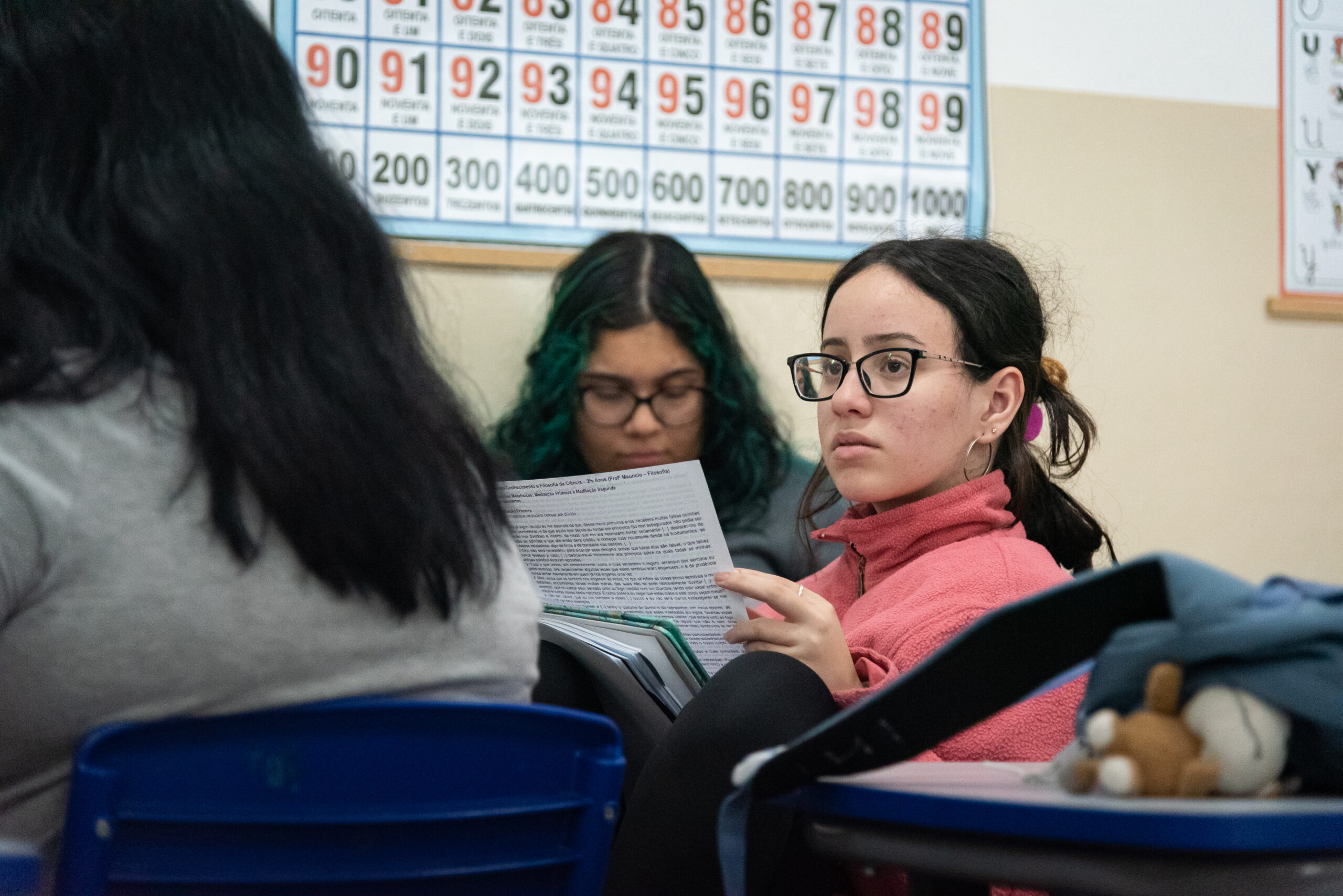Fotografia de estudantes do Ensino Médio no centro da foto uma estudantes de blusa rosa e óculos segura uma folha de texto.