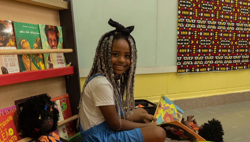 Fotografia de uma menina negra olhando para a câmera e sorrindo. Ela está sentada no chão e com um livro em suas mãos. Ao fundo uma estante com variedade de livros para as ações antirracistas.