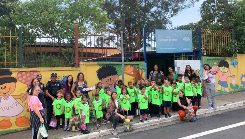 Fotografia de crianças com colete verde e alguns adultos educadores em frente a escola no prjeto sobre alimentação saudável.