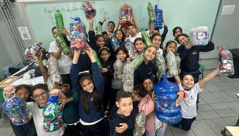 Fotografia de vários estudantes com os braços para cima segurando garrafas de plátstico com milhares de lacres de latinhas dentro na ação do projeto "Educa Verde" da DRE Penha.