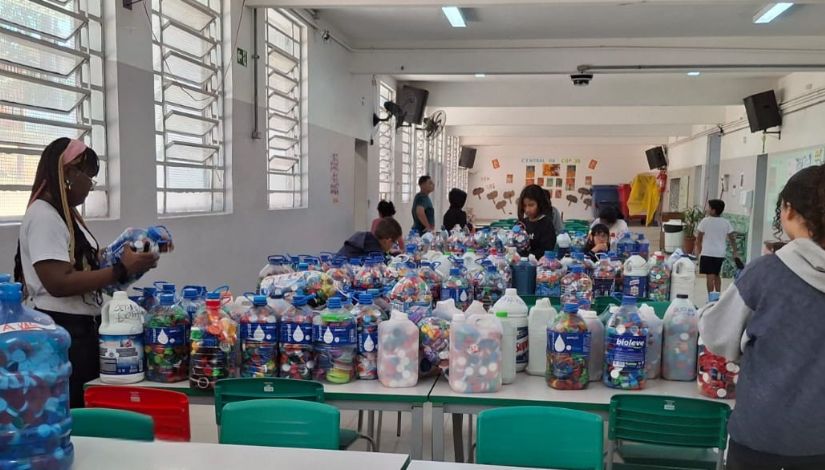 Fotografia de um grande espaço com mesas e sobre elas inúmeras garrafas de plástico com tampinhas também de plástico dentro e alguns estudantes organizando as mesmas na ação do projeto "Educa Verde" da DRE Penha.