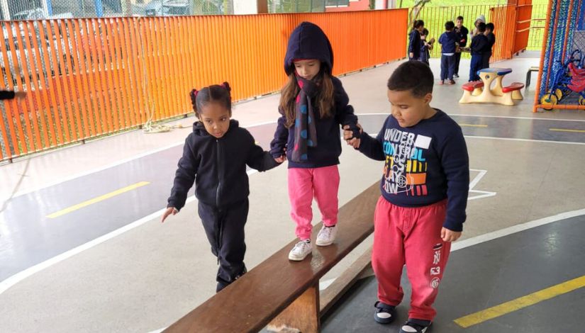 Uma criança andando sobre um banco longo de madeira com a auxilio de uma amigo e uma amiga que seguram suas mão no projeto.