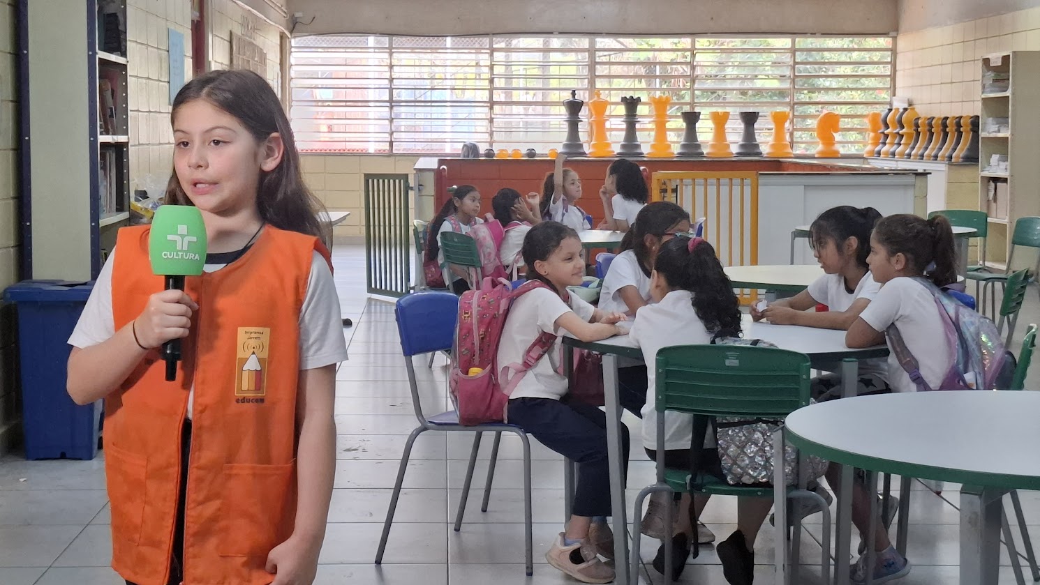 Estudante do Imprensa Jovem com colete laranja apresentando programa com microfone da TV Cultura ao fundo crianças em uma mesa conversando