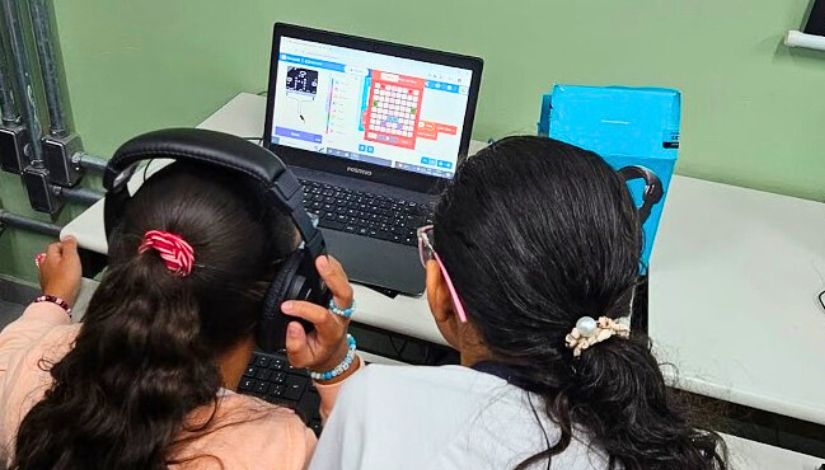 Fotografia mostra duas estudantes de costas para a fotografia e olhando para um programa digital no computador de mesa.