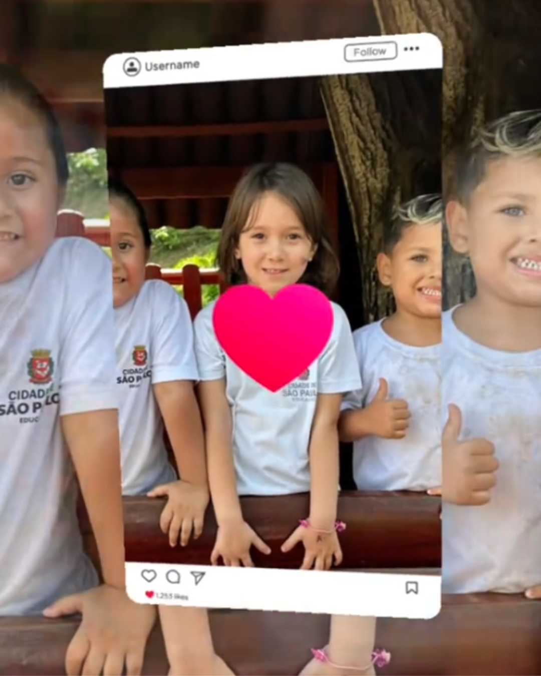 Imagem de três crianças pequenas, em ambiente externo, apoiadas em uma estrutura de madeira, como um parquinho. Elas vestem camisetas brancas com o brasão da Prefeitura de São Paulo. Ao centro, uma criança com cabelos castanhos médios sorri para a câmera, com um coração rosa sobreposto na região do peito. À direita, outra criança sorri e faz sinal de positivo com o polegar. À esquerda, parte do rosto e do corpo de uma terceira criança também aparece. A imagem está enquadrada como se fosse uma postagem de rede social, com elementos gráficos simulando uma interface, como nome de usuário e ícones de interação. Ao fundo, há árvores e uma estrutura de madeira, indicando um espaço ao ar livre.
