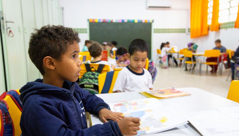 Imagem de uma sala de aula com crianças pequenas sentadas em mesas individuais. Em primeiro plano, um menino com cabelo curto e encaracolado, usando casaco azul, está sentado de perfil, observando o ambiente enquanto segura um livro aberto sobre a mesa. Ao lado, outra criança lê atentamente. Ao fundo, outras crianças estão distribuídas pela sala, envolvidas em atividades de leitura e escrita. O ambiente é bem iluminado, com cadeiras coloridas, quadro ao fundo e cortinas amarelas nas janelas, transmitindo um cenário de aprendizagem acolhedor e organizado.