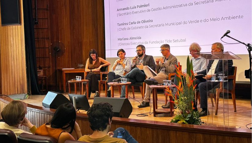 Painel com seis participantes sentados em cadeiras sobre um palco, durante um evento, debatendo temas relacionados à gestão pública. Ao fundo, há uma tela com os nomes dos palestrantes, enquanto o público acompanha a conversa da plateia.