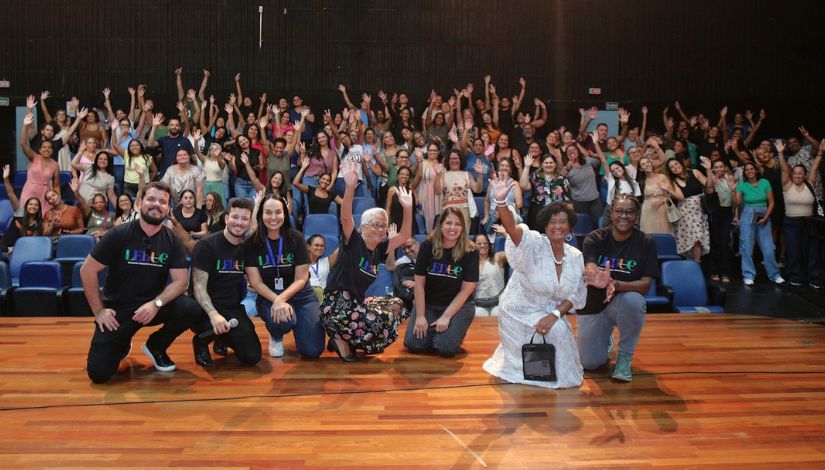 Foto de um grande grupo de pessoas reunidas em um auditório, com participantes em pé nas arquibancadas ao fundo e um grupo menor ajoelhado na frente do palco. Todos sorriem e acenam para a câmera, transmitindo um clima de celebração e participação coletiva.