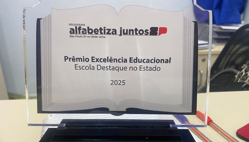 Placa de premiação em acrílico com o texto “Prêmio Excelência Educacional – Escola Destaque no Estado 2025”, do programa Alfabetiza Juntos SP. O troféu está apoiado sobre uma base, em ambiente interno.