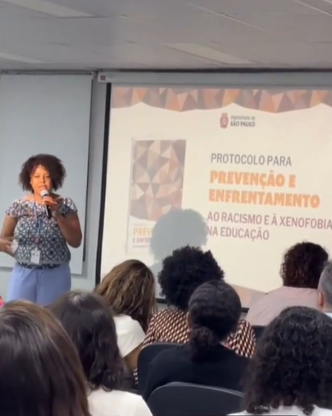 Mulher com microfone na mão ao lado do telão palestrando para as demais pessoas sentadas à frente de telão.