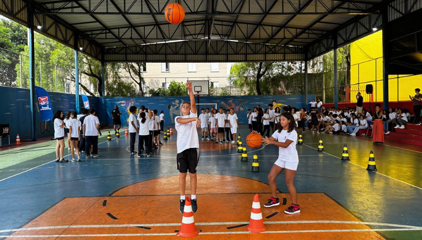 Estudantes com uniforme da prefeitura praticam basquete em uma quadra
