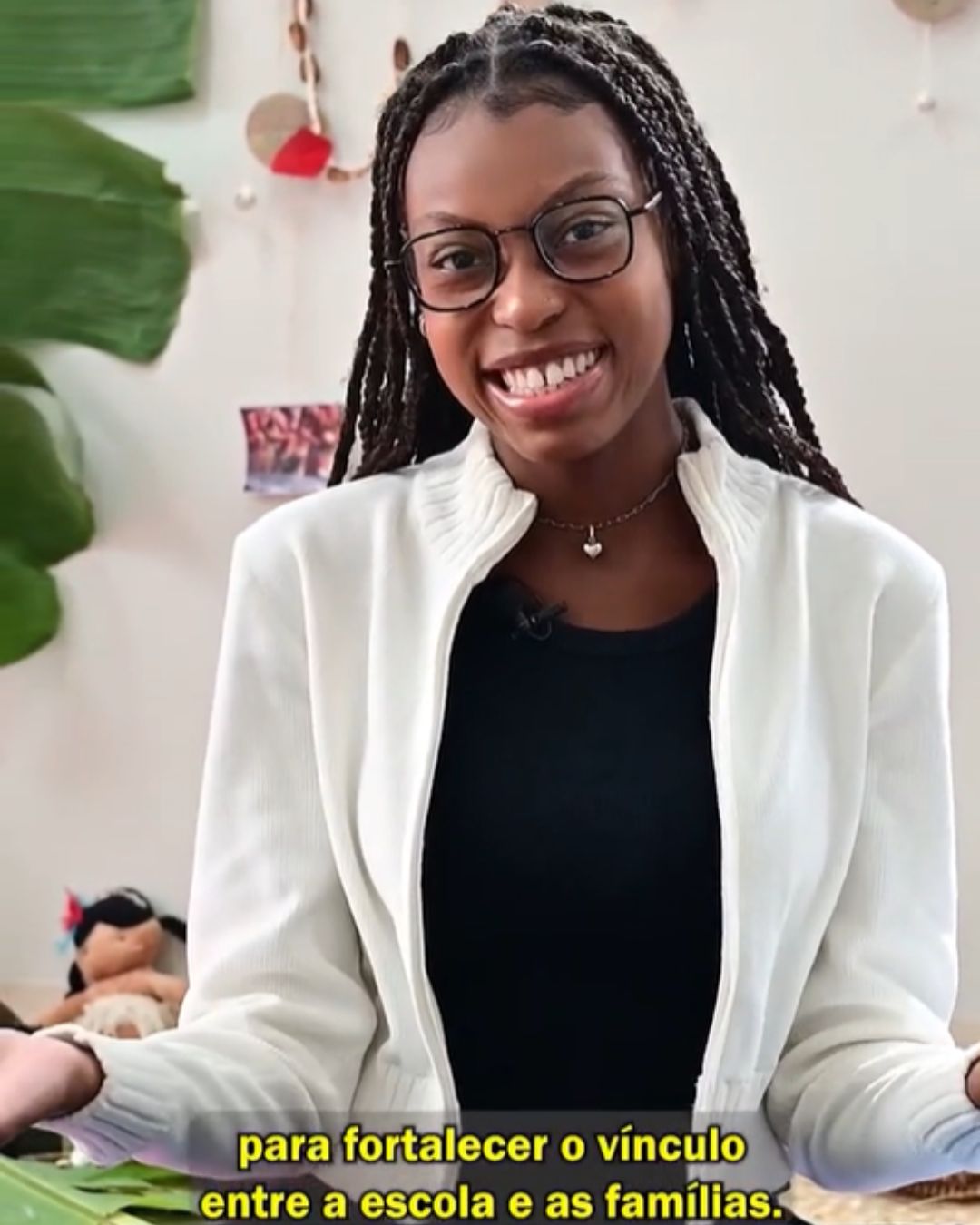Menina, de óculos e cabelos trançados, sorri enquanto fala para a câmera em um ambiente interno com plantas e objetos ao fundo.