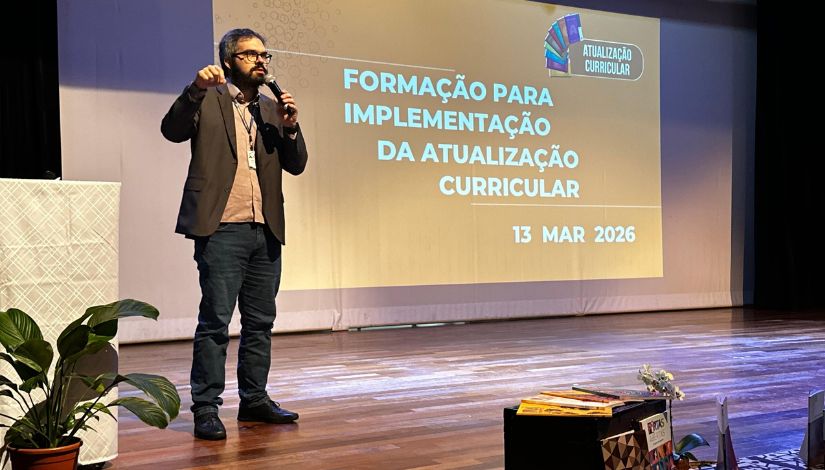 Fotografia de um homem em pé no placo falando ao microfone e ao fundo projetado em um teláo o texto 