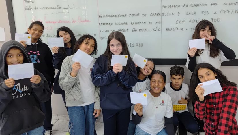 Fotografia com várias meninas e meninos. Eles estão em frente a uma lousa de vidro e seguram envelopes de cartas.