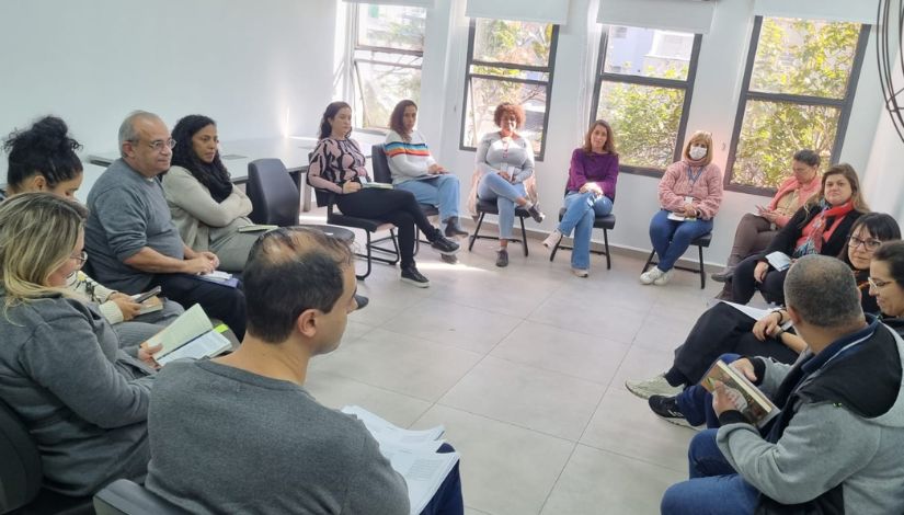 Fotografia de quinze adultos sentados em círculo com livros nas mãos durante o clube de leitura.