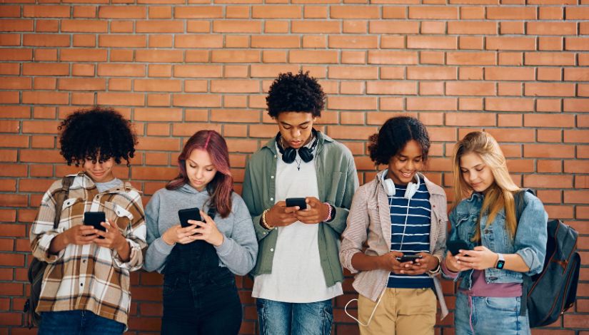Fotografia mostra cinco jovens segurando celulares e olhando para as respectivas telas. Ao fundo parede de tijolos a vista.