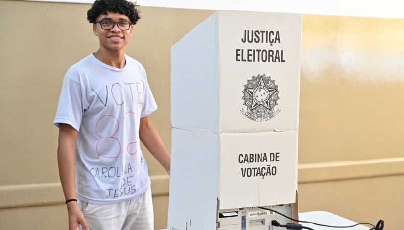 Imagem de um jovem sorridente, em ambiente interno, ao lado de uma cabine de votação. Ele usa óculos, camiseta clara com a palavra “VOTE” e o número “8” escritos à mão, além de um nome abaixo, sugerindo apoio a uma candidatura. Ao lado, há uma estrutura de papelão branca com as inscrições “Justiça Eleitoral” e “Cabina de Votação”, com o símbolo oficial ao centro. A cabine está posicionada sobre uma mesa, onde também é possível ver parte de um equipamento eletrônico. O cenário remete a um processo de votação escolar, como eleições de grêmio estudantil.