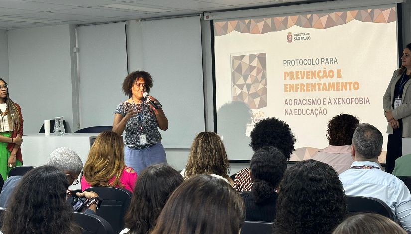 Fotografia de uma mulher negar ao centro em frente ao público do auditório, segurando o microfone. Ao fundo projetado no telão o texto “Protocolo para Prevenção e Enfrentamento ao Racismo e à Xenofobia na Educação”.