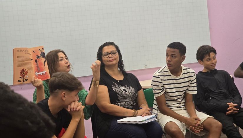 Fotografia e uma mulher segurando um livro e mostrando para os adolescentes na roda de conversa.