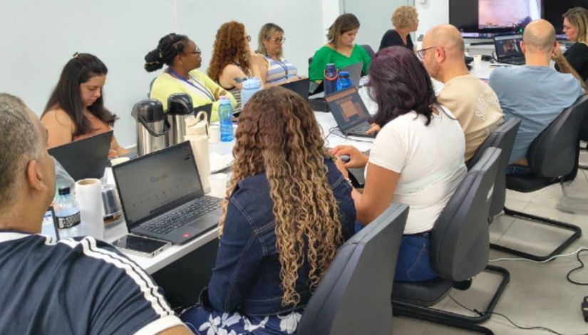 Fotografia de vários professores sentados ao redor da mesa com seus computadores portáteis.